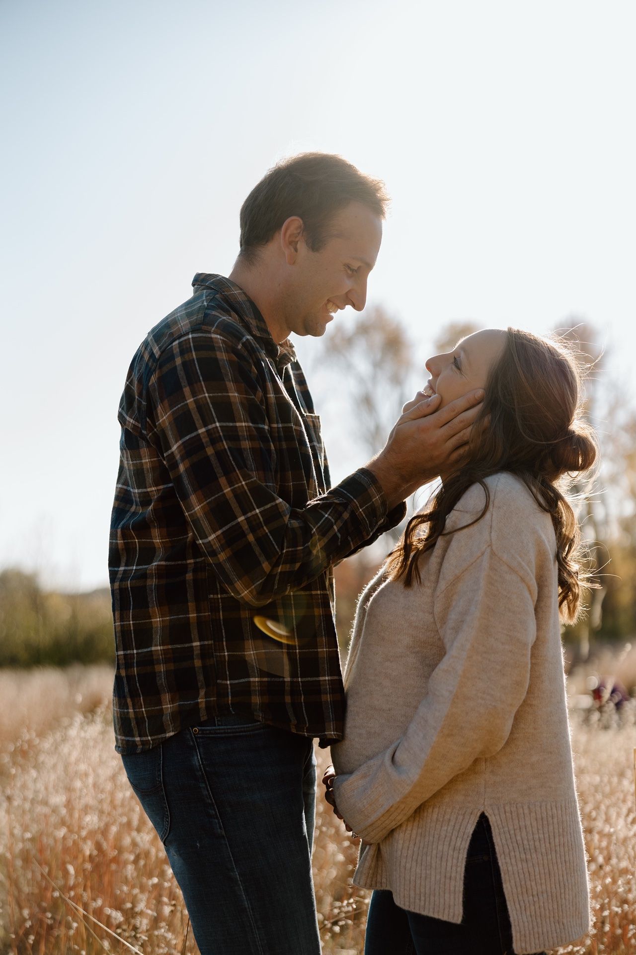 A pregnant couple shares a tender moment in a sunlit field, the man gently cupping the woman's face as she laughs up at him.