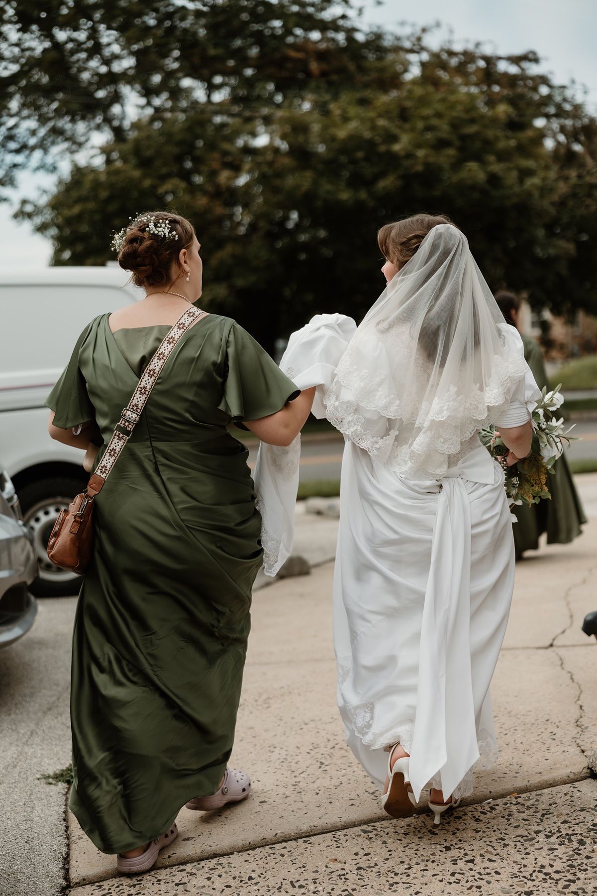 Bride in white gown and veil walks with bridesmaid in olive green dress along sidewalk on wedding day.