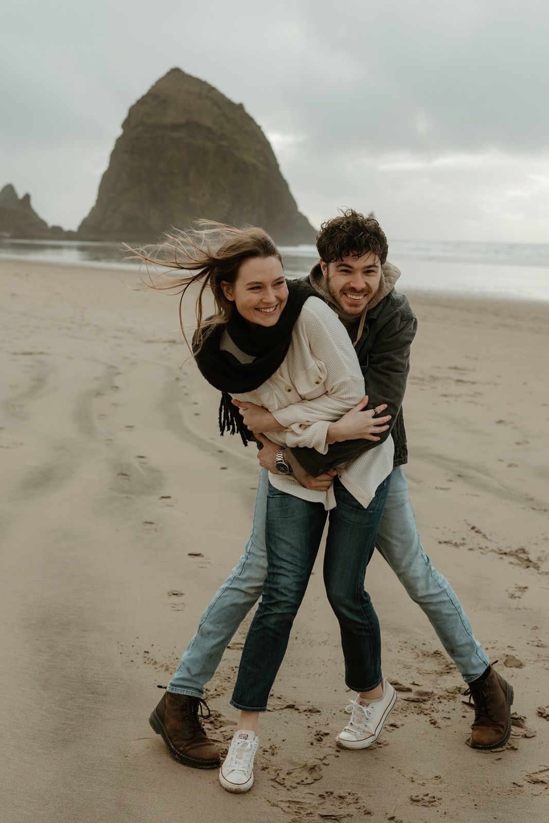 A joyful couple embraces and laughs on a sandy beach with Haystack Rock in the background on a cloudy day.