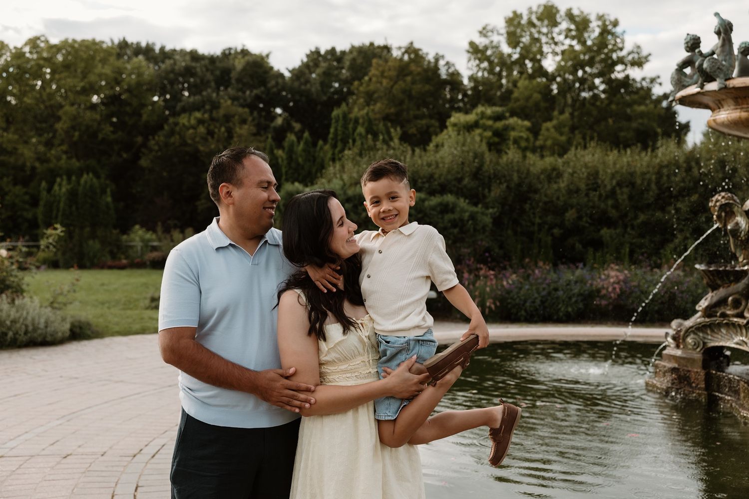 Family of three smiling by a garden fountain, parents holding young boy outdoors.