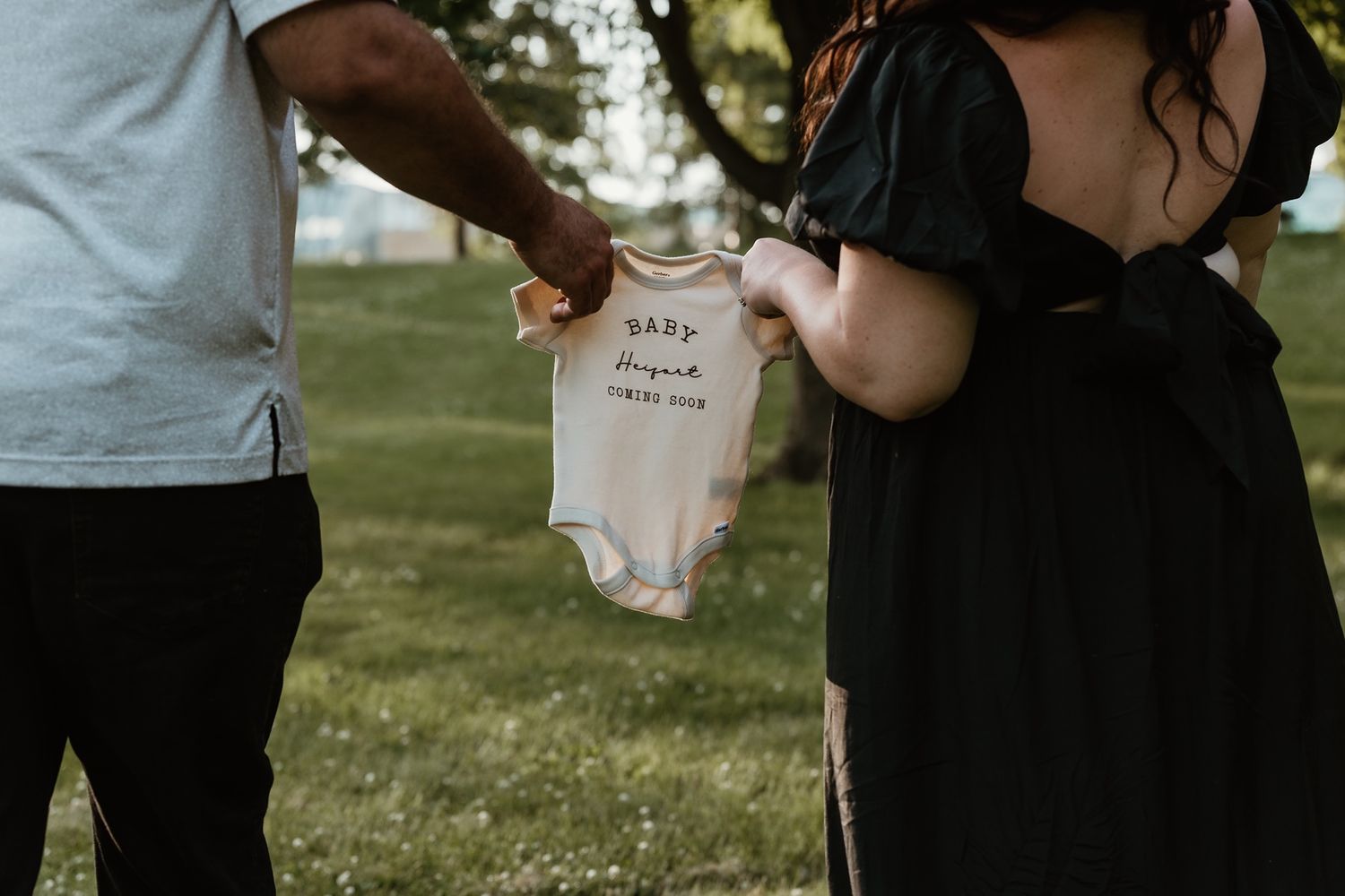 Couple holding a baby onesie outdoors, announcing pregnancy with a 'Baby Coming Soon' bodysuit in a park.