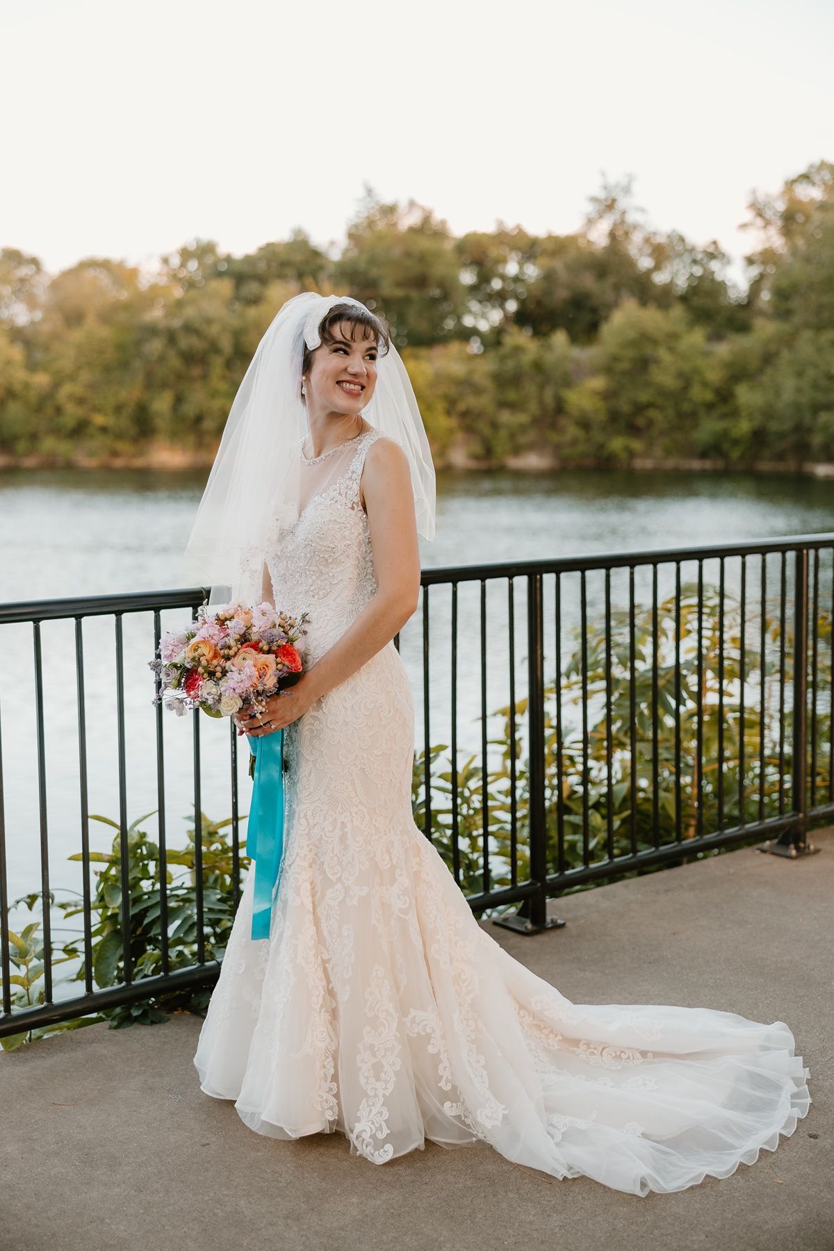 Bride in lace mermaid gown and veil holds colorful bouquet with teal ribbon, posing on outdoor terrace near lake.