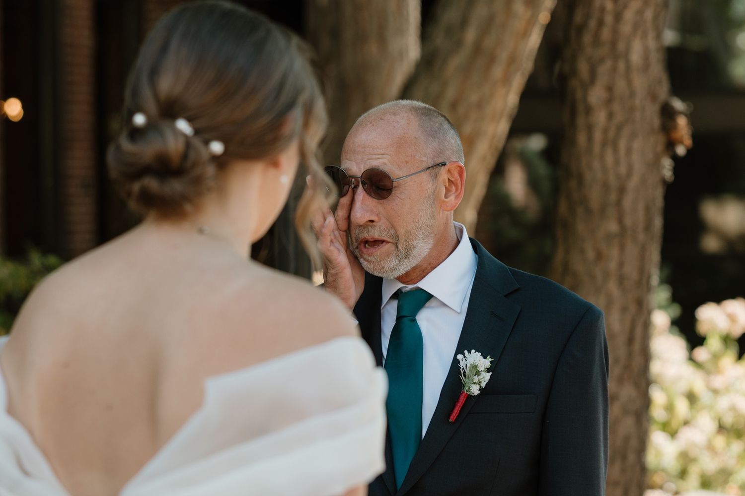 Emotional father wiping tears seeing bride daughter in white wedding dress outdoors among trees.