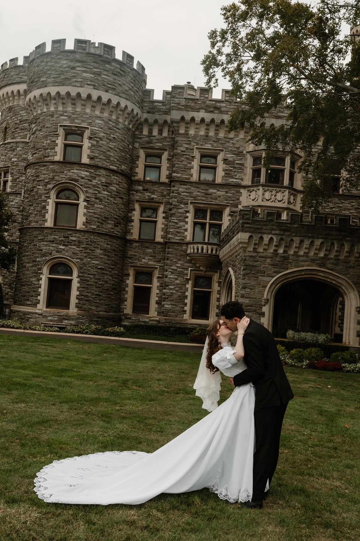 Bride and groom sharing a romantic dip kiss on green lawn in front of a grand stone castle wedding venue.
