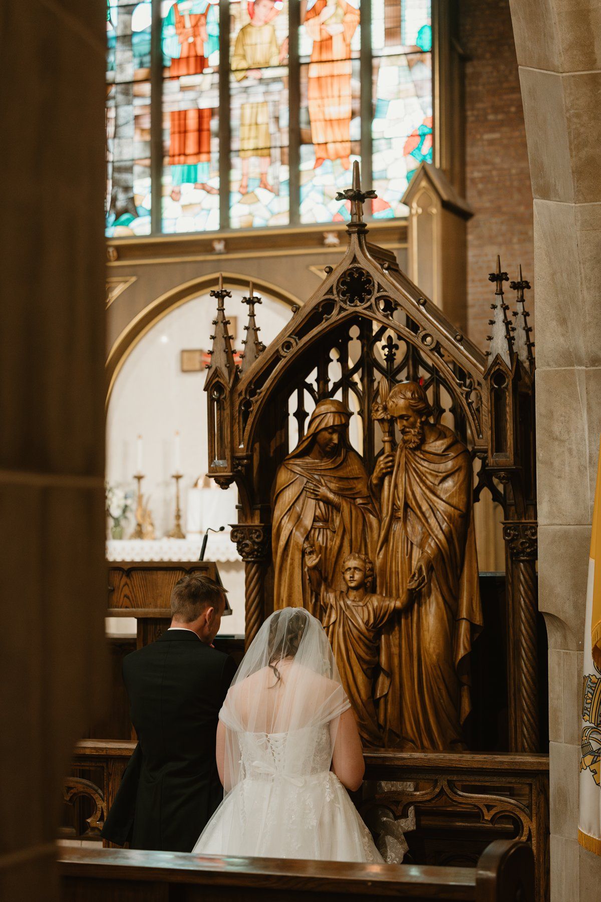 A bride in a white dress stands near ornate wooden church pews with carved decorative details and stained glass windows.