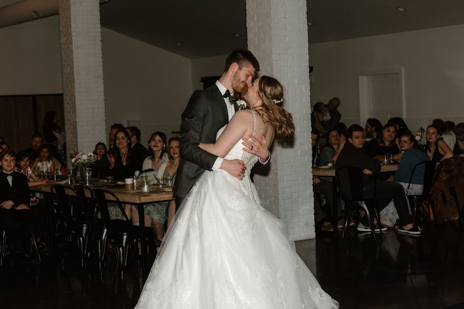 Wedding couple dancing surrounded by guests in a dimly lit reception hall with white decor.