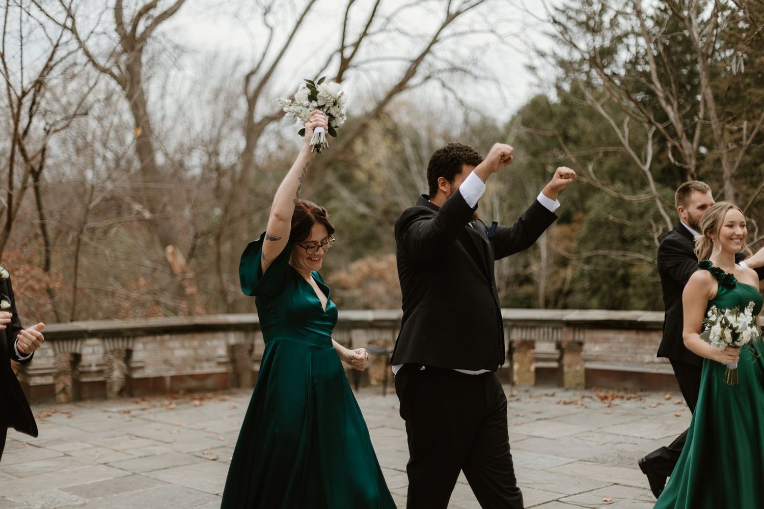 Wedding party members dance and celebrate outdoors with raised arms in emerald green dresses.