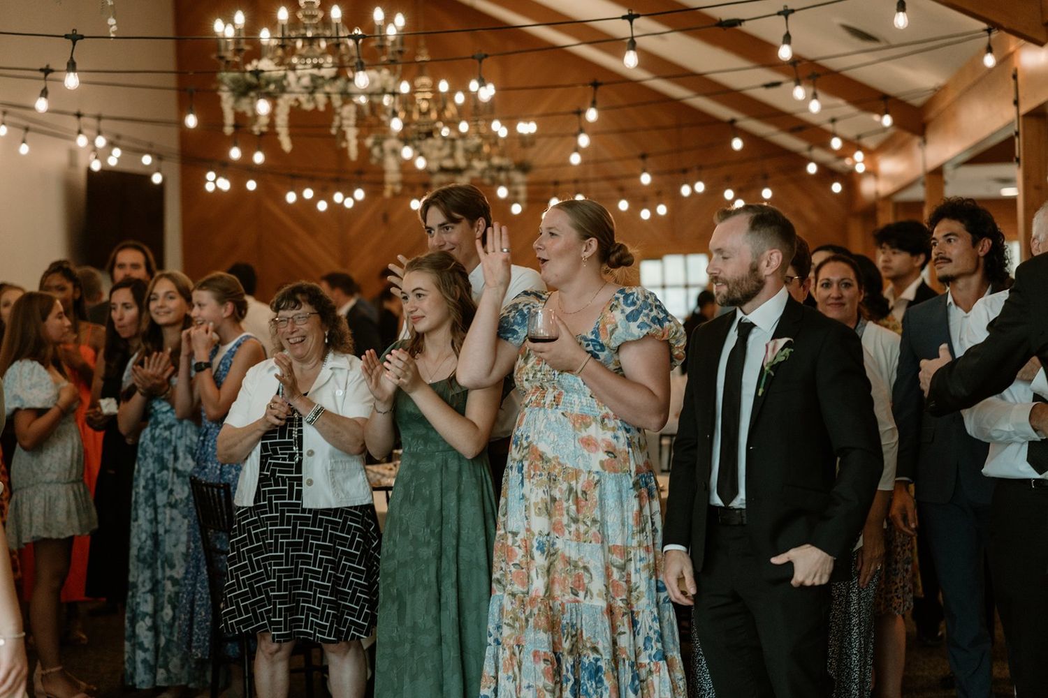Wedding guests dancing and celebrating on a dance floor under glowing chandeliers at an indoor reception venue.