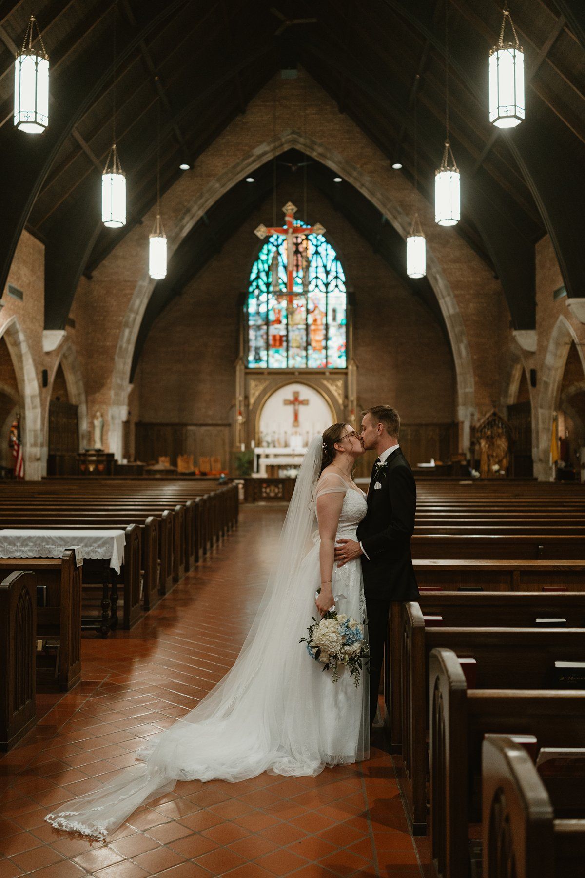 A couple shares an intimate moment in a stone church with stained glass windows and wooden pews.
