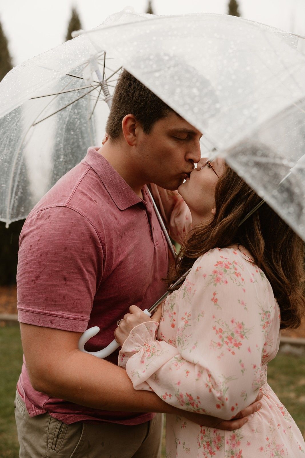 A romantic couple kisses beneath a clear umbrella in the rain while wearing pink and floral outfits.