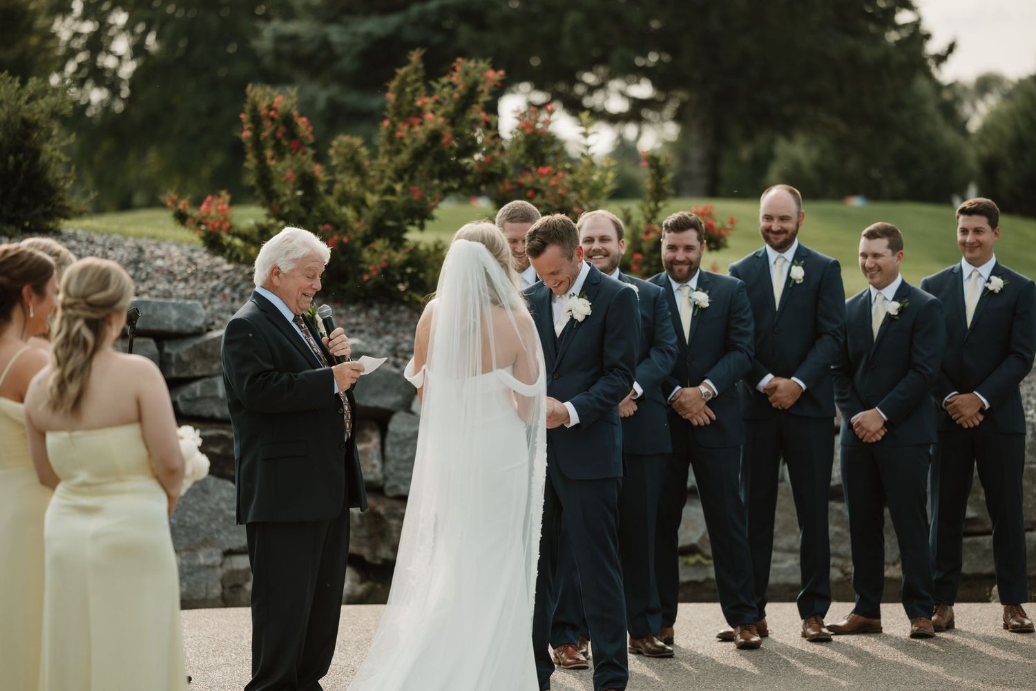 Wedding party in navy suits and light dresses poses outdoors during sunset for formal group photos.