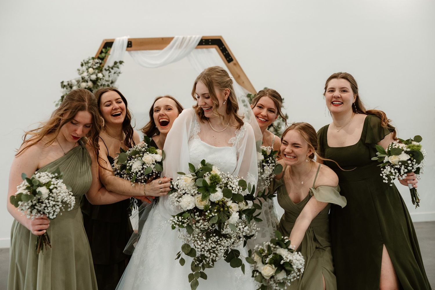 Bridesmaids in black dresses and bride in white laugh together while holding white floral bouquets at outdoor wedding.