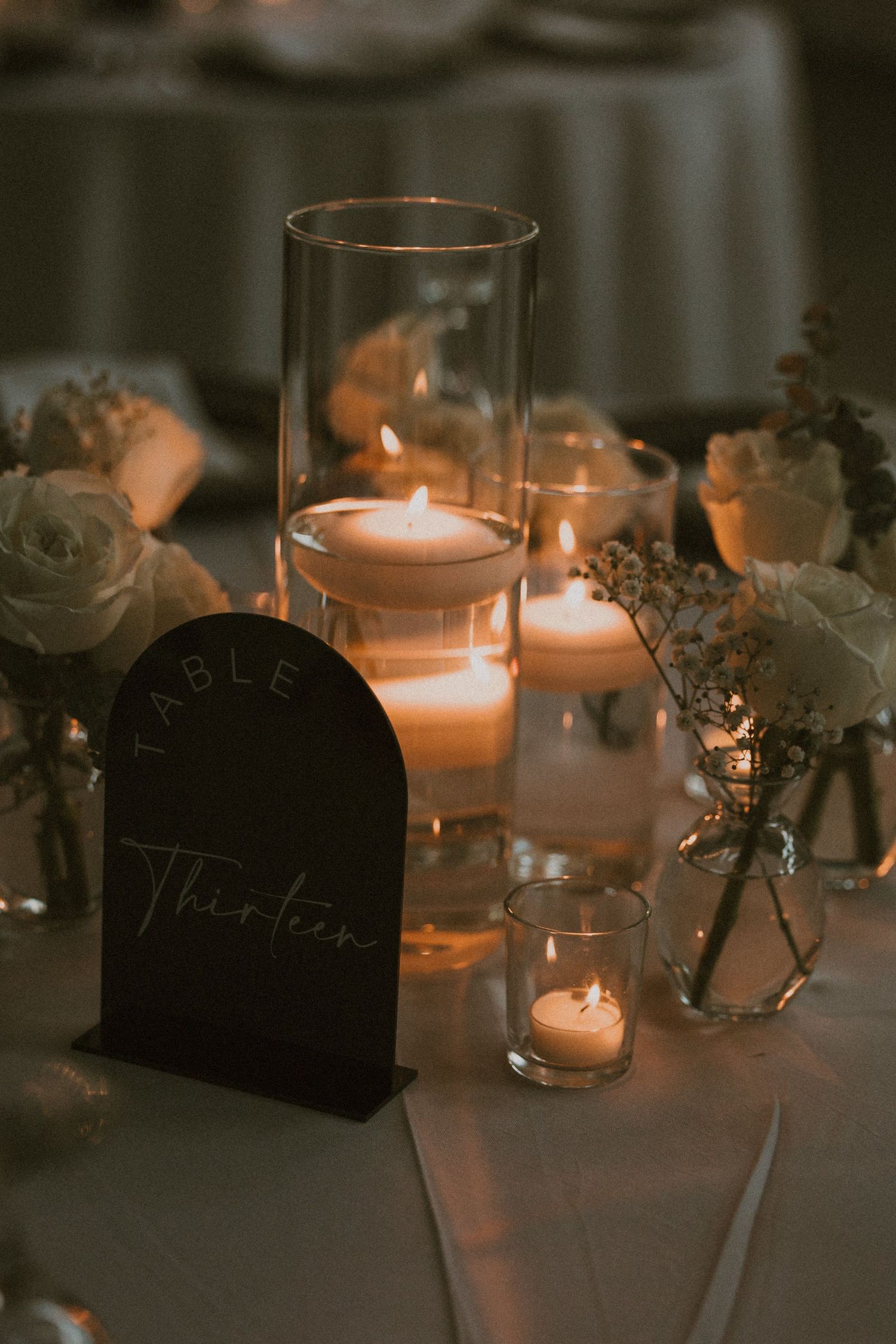 Romantic candlelit table setting with glass votives, roses and a black table number holder creating an intimate ambiance.