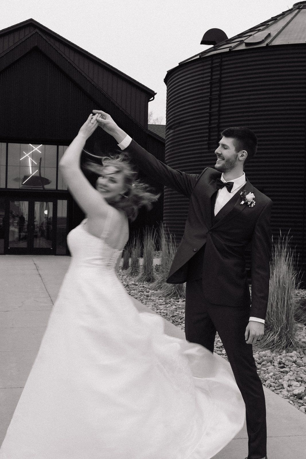 A bride and groom share a playful dance moment against a backdrop of dark metal grain silos.