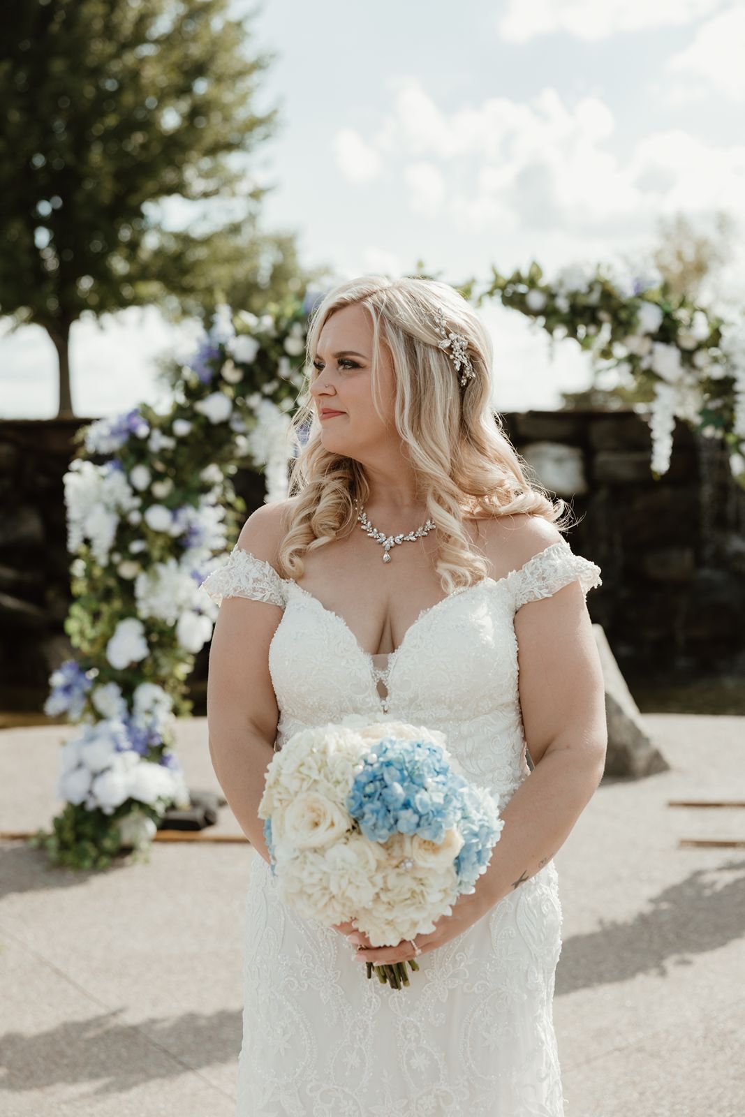 A bride in a lace wedding dress poses with her white and blue hydrangea bouquet on her wedding day.