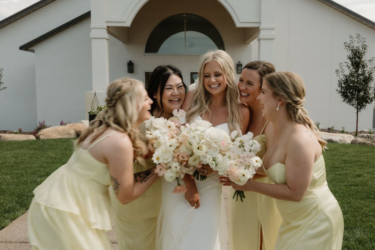 Bridesmaids in pale yellow dresses share a candid laughing moment while holding bouquets at an outdoor wedding.