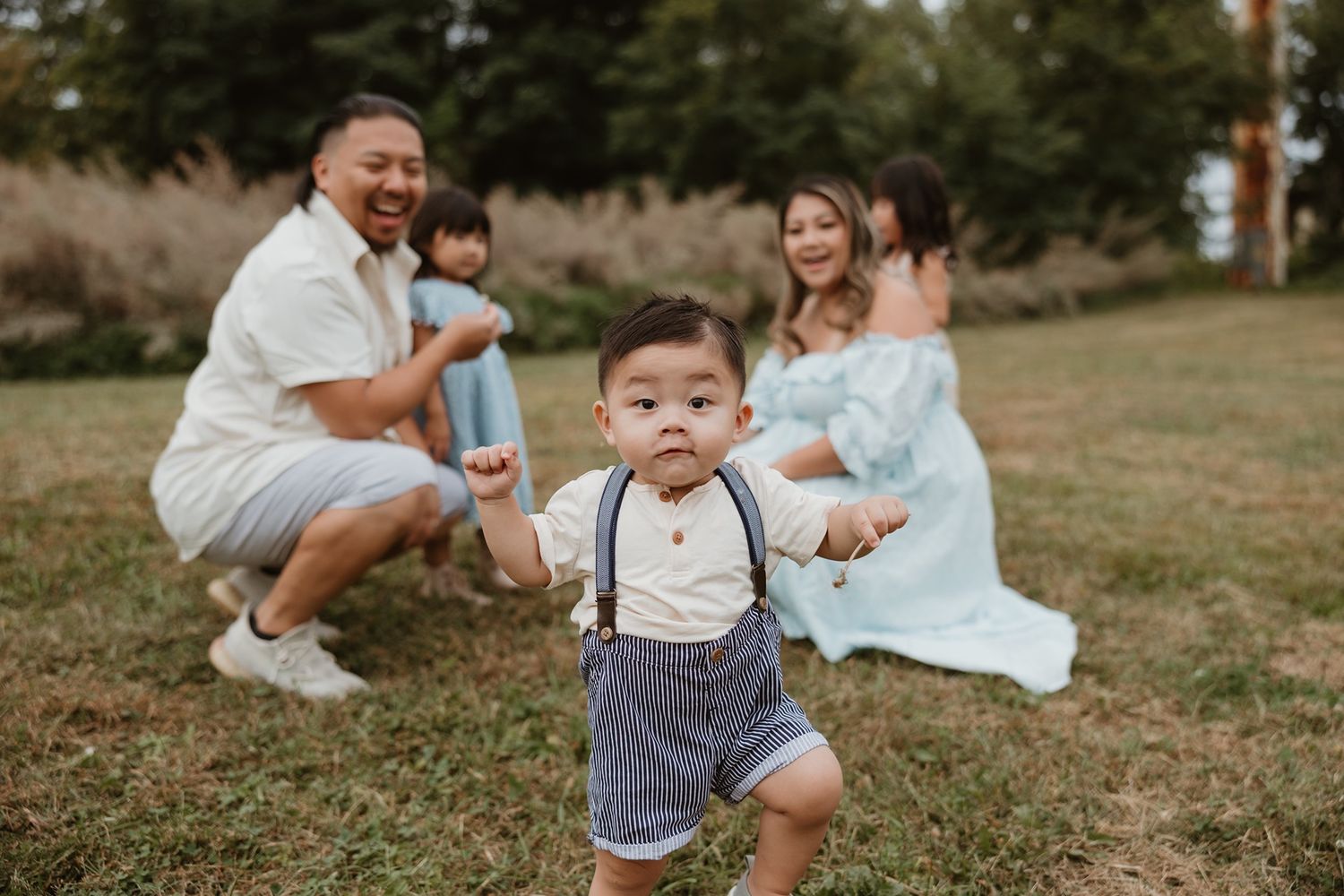 A joyful family poses outdoors, with a toddler boy in suspenders walking toward the camera while parents and sibling smile.