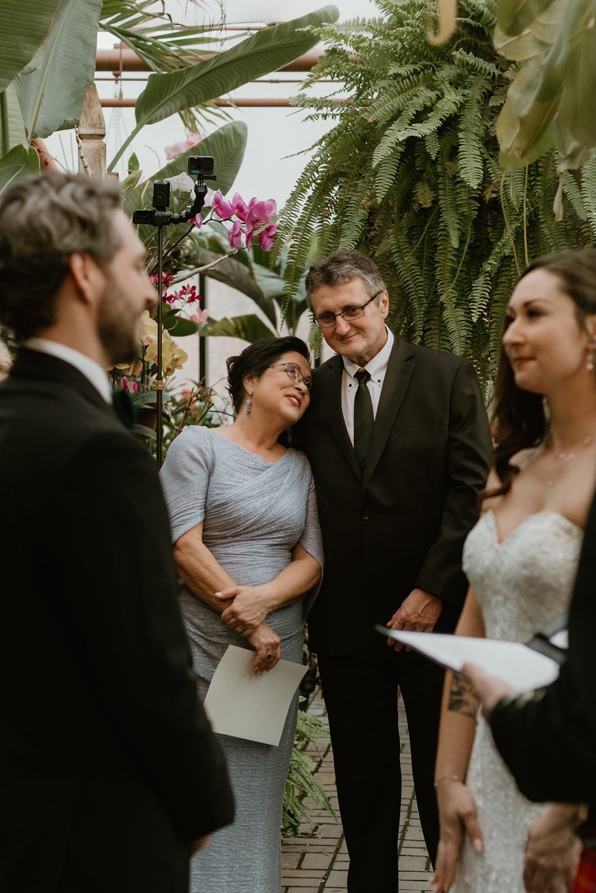 Couple smiles during intimate wedding ceremony surrounded by tropical greenery and orchids in a lush botanical venue.