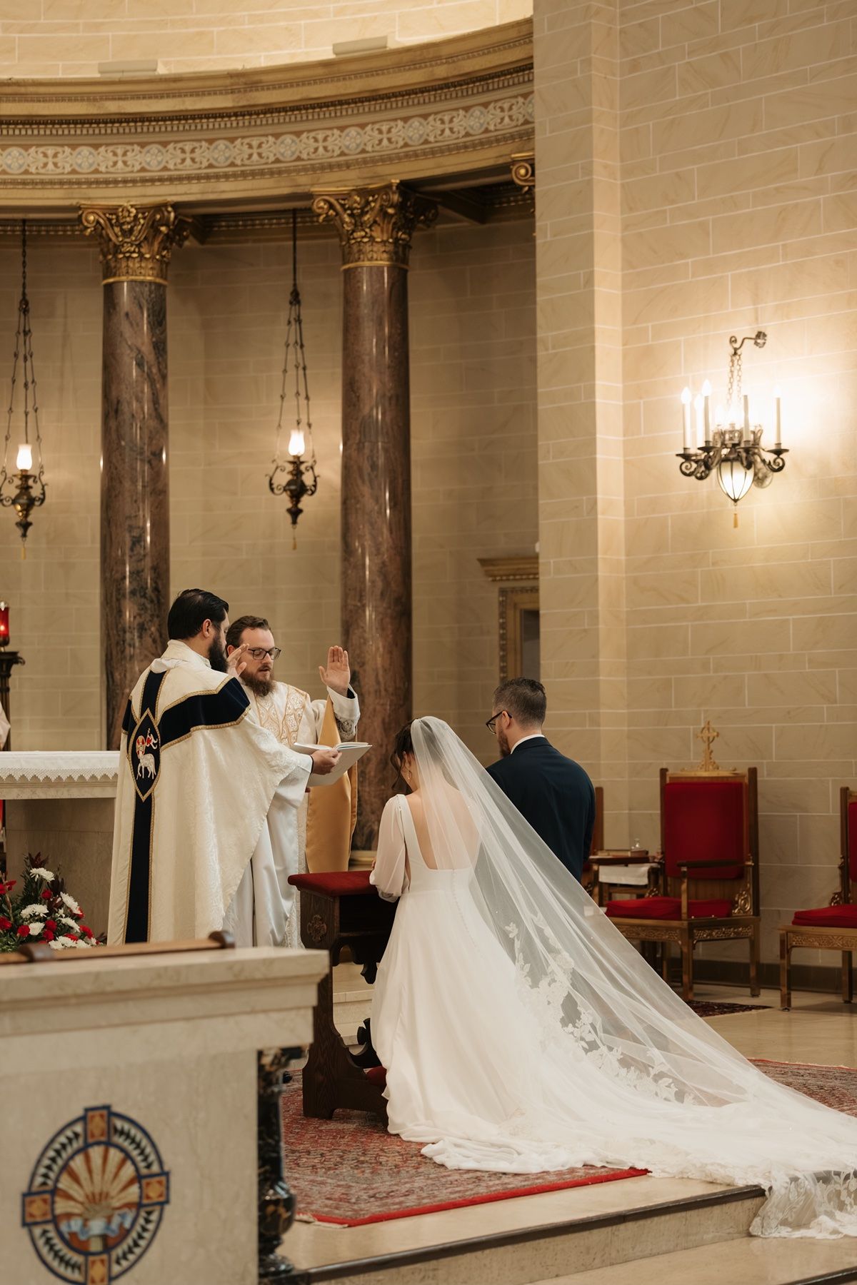 Bride and groom kneel at church altar during wedding ceremony with priest officiating in ornate chapel.