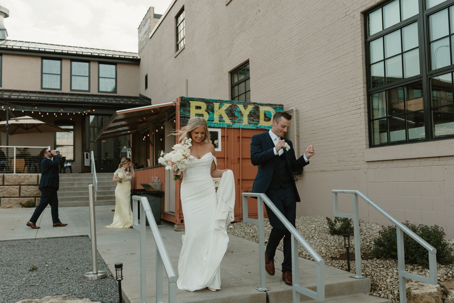 A wedding party walks down a paved path between modern buildings on a sunny day.