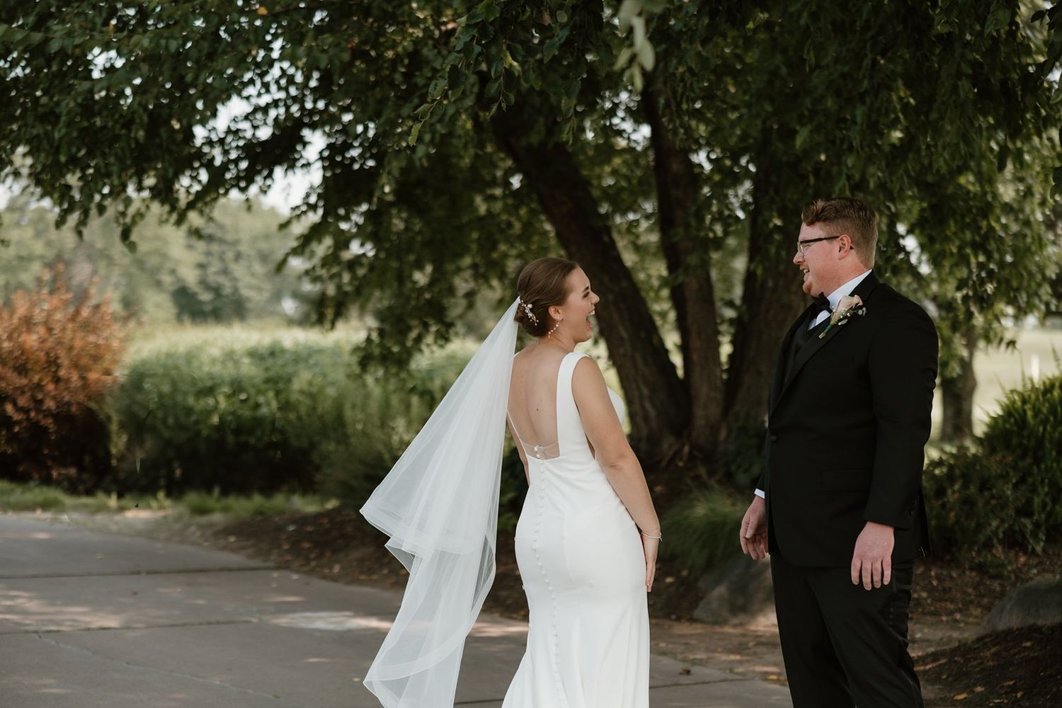 A couple shares a romantic moment on their wedding day under a shady tree on a garden path.