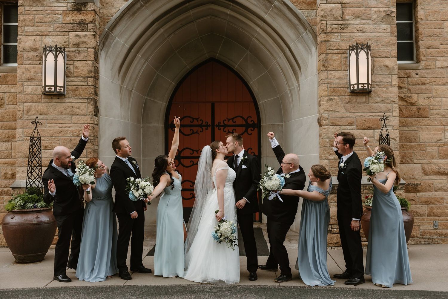 A bridal party wearing dusty blue bridesmaid dresses and formal attire poses in an archway of a stone building.