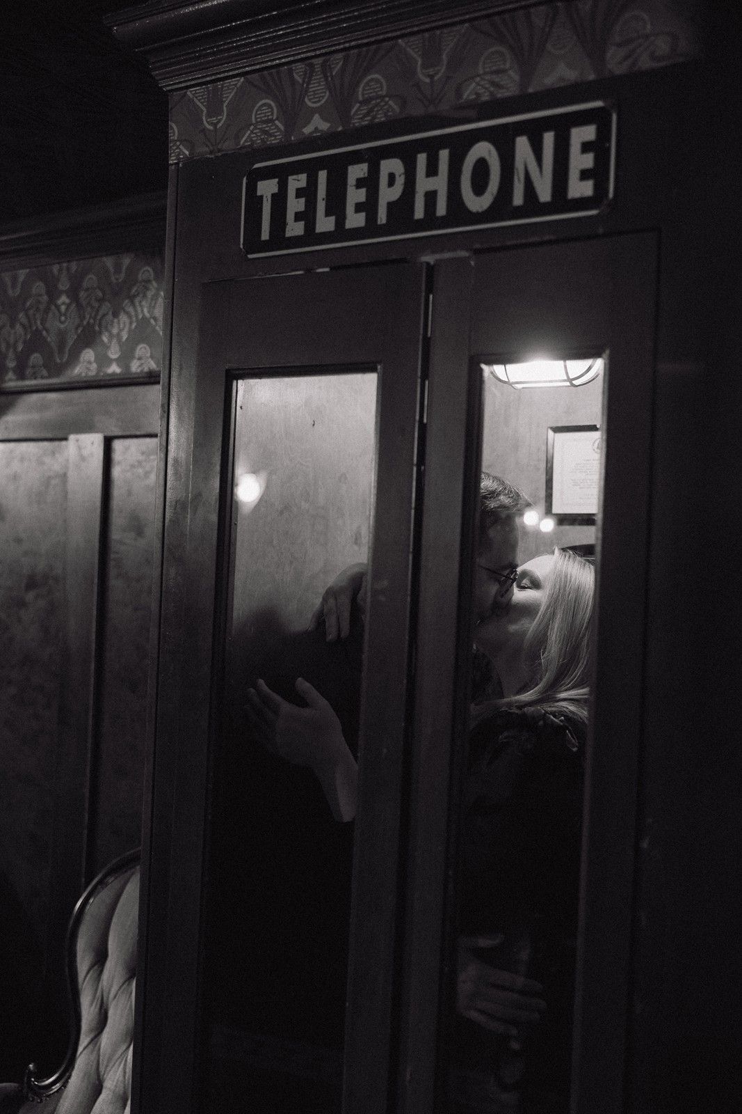 A dark moody black and white photograph of a vintage telephone booth with silhouettes visible through frosted glass.