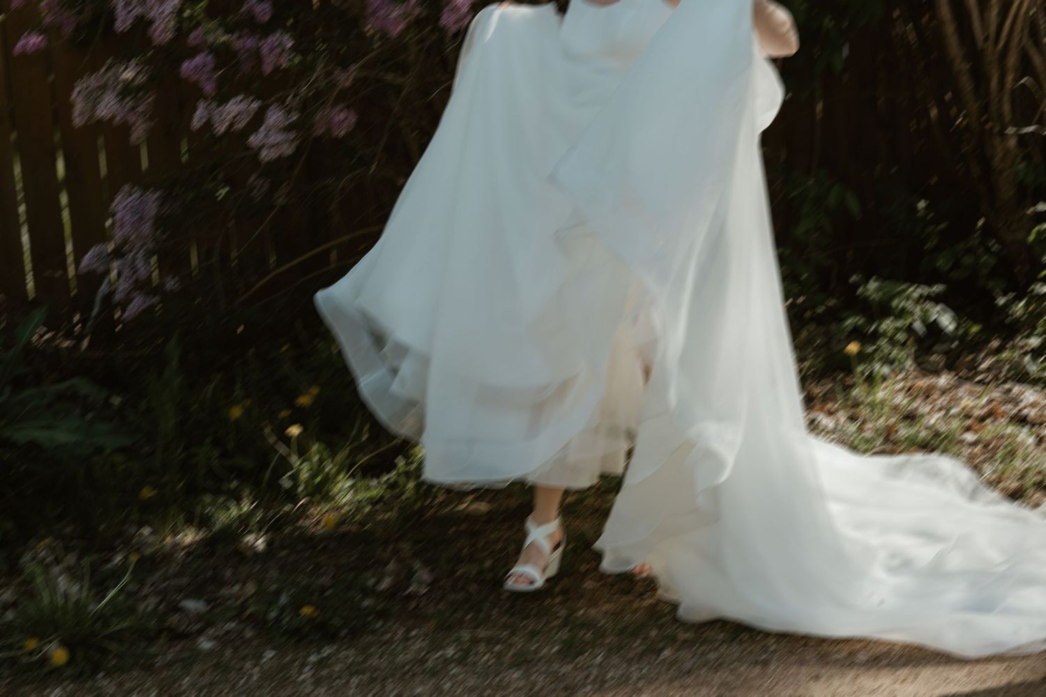 Bride in flowing white wedding gown with train walks through garden path surrounded by purple blooming flowers.