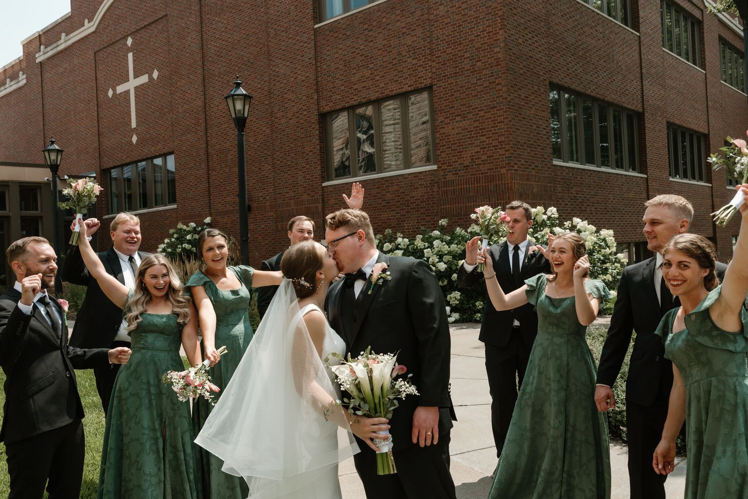 Wedding party celebrates with arms raised in joyful dance moves on outdoor garden path during reception.