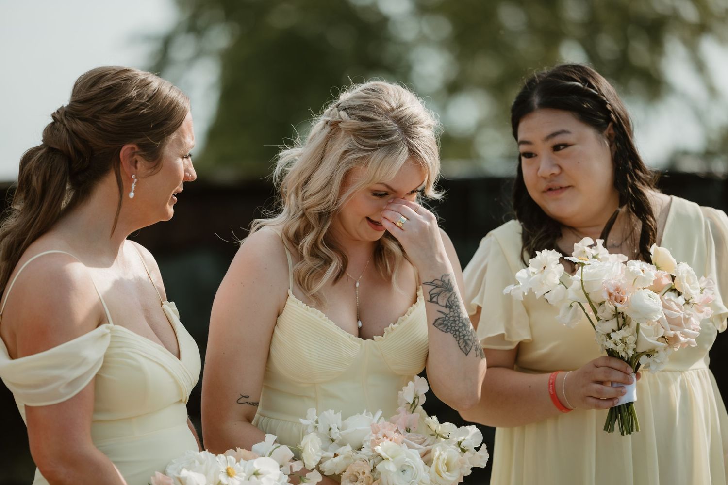 A row of bridesmaids in cream-colored dresses hold white bouquets during an outdoor wedding ceremony.