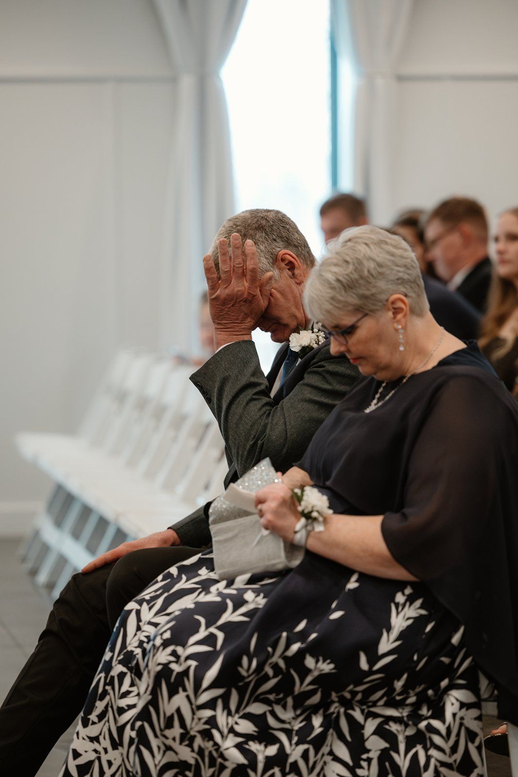 An emotional moment during a wedding ceremony as attendees bow their heads in prayer while seated on white chairs.