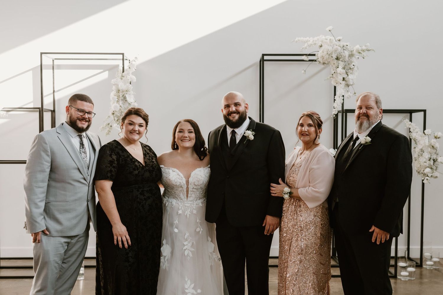 A wedding party poses together at a modern venue decorated with white floral arrangements and metallic frames.