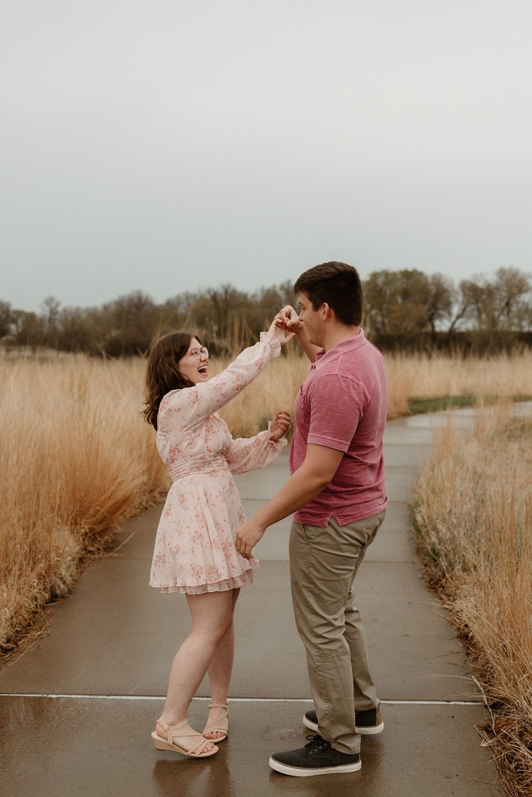 A couple share a playful moment on a pathway surrounded by tall brown grass on an overcast day.