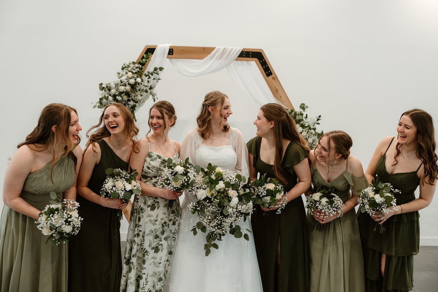 A wedding party in olive green and black dresses laughs together while holding white floral bouquets.
