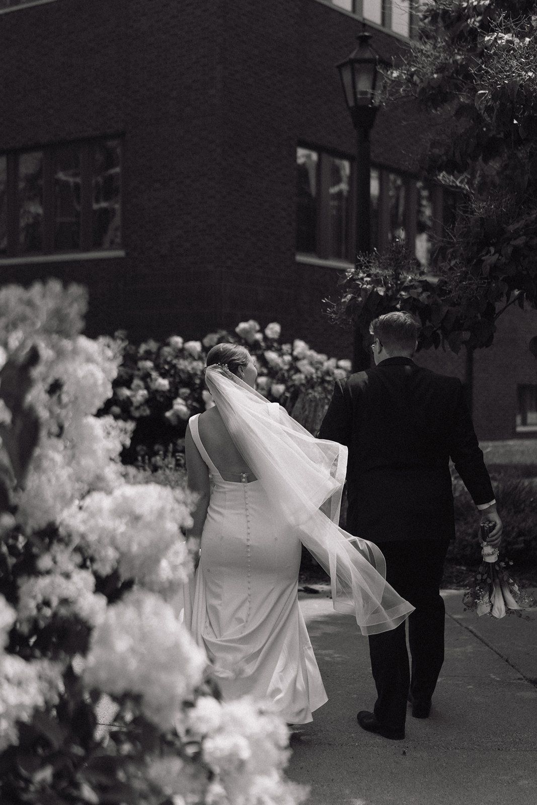A black and white wedding photo showing a couple walking past decorative white flowers with a flowing veil in motion.