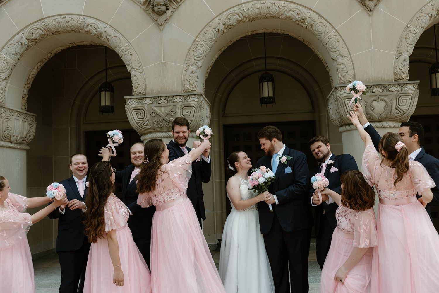 Wedding party with bridesmaids in pink dresses and groomsmen in black suits pose together under ornate architecture.