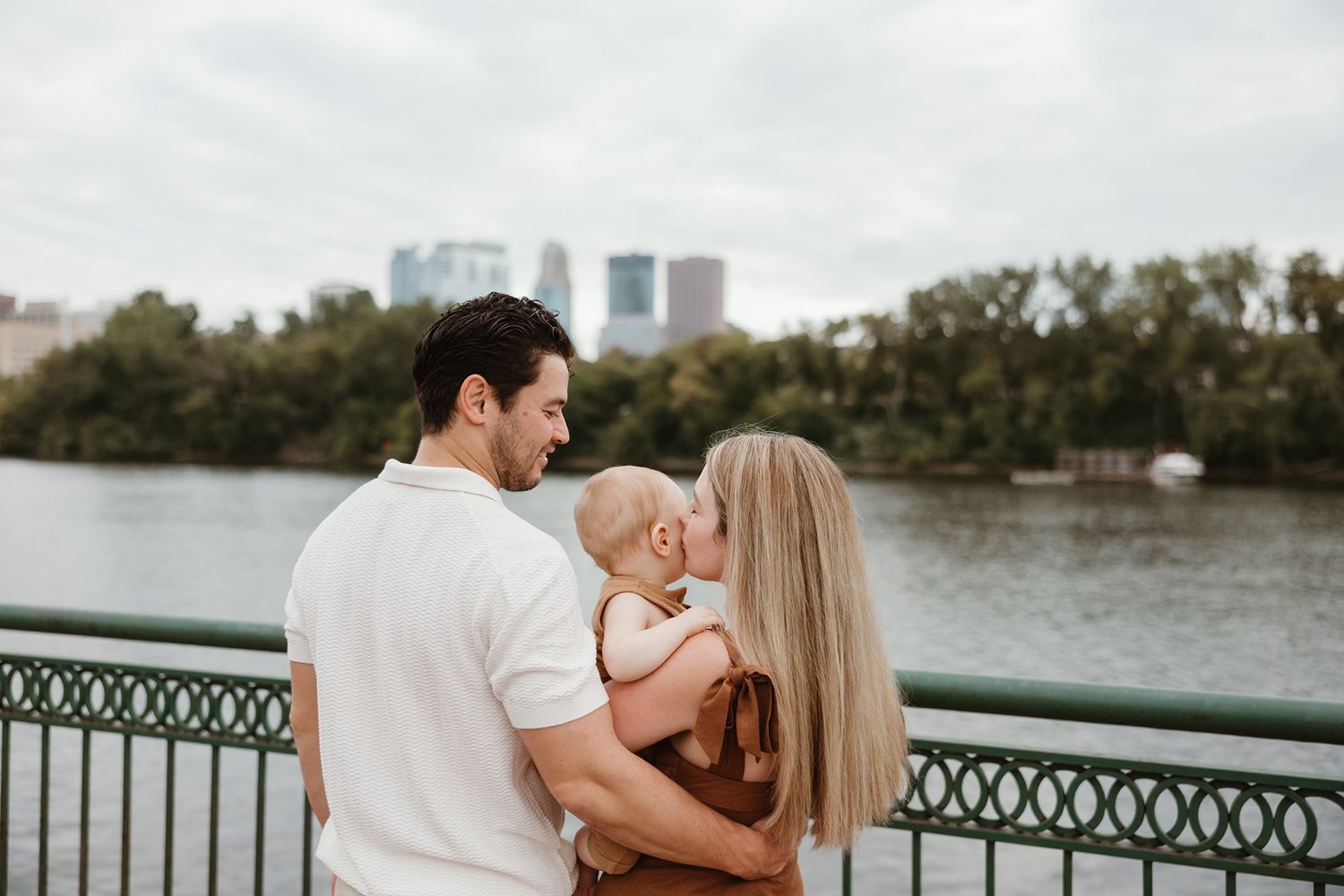 A couple kisses their baby on a bridge overlooking a river with a city skyline and trees in the background.