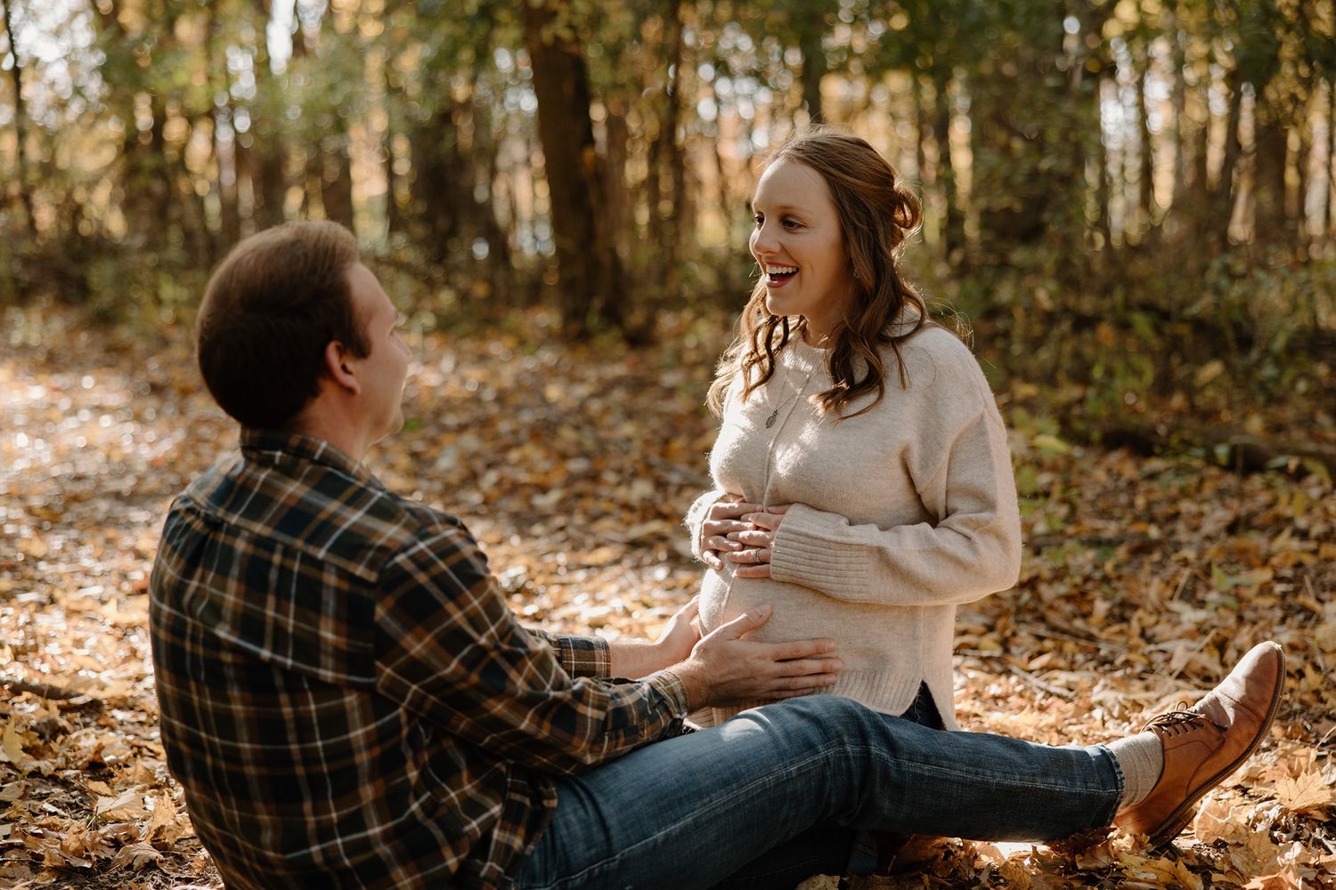 Couple sitting outdoors in autumn forest, pregnant woman smiling and holding baby bump during maternity photo session.