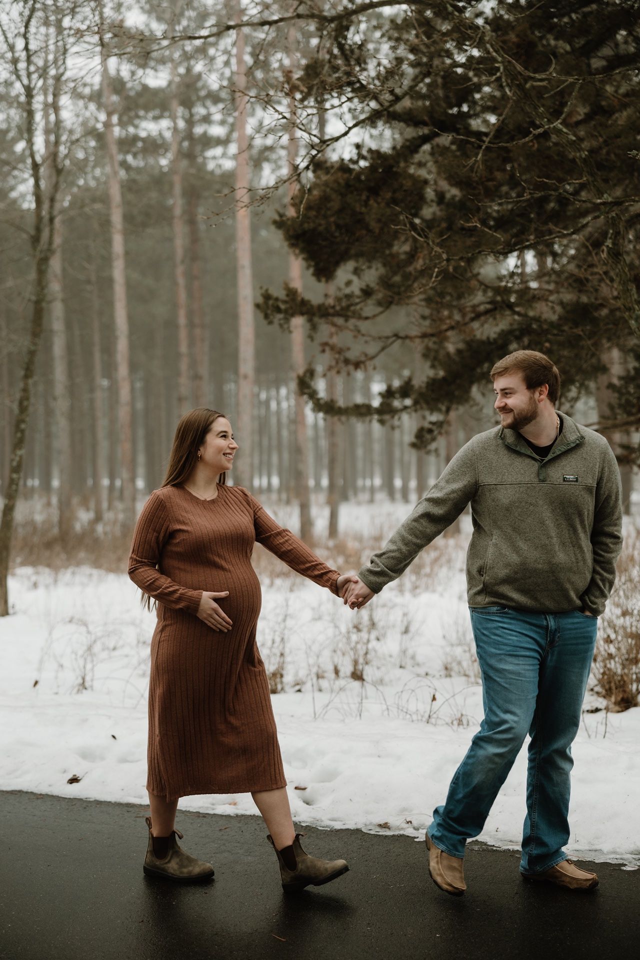 Pregnant couple holding hands and smiling on a snowy winter path surrounded by trees, woman in brown dress touching baby bump.