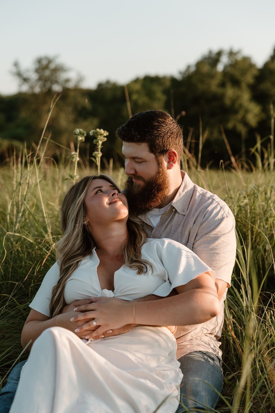 A couple sits in a sunlit field, the bearded man embracing the woman in a white dress as they gaze at each other.