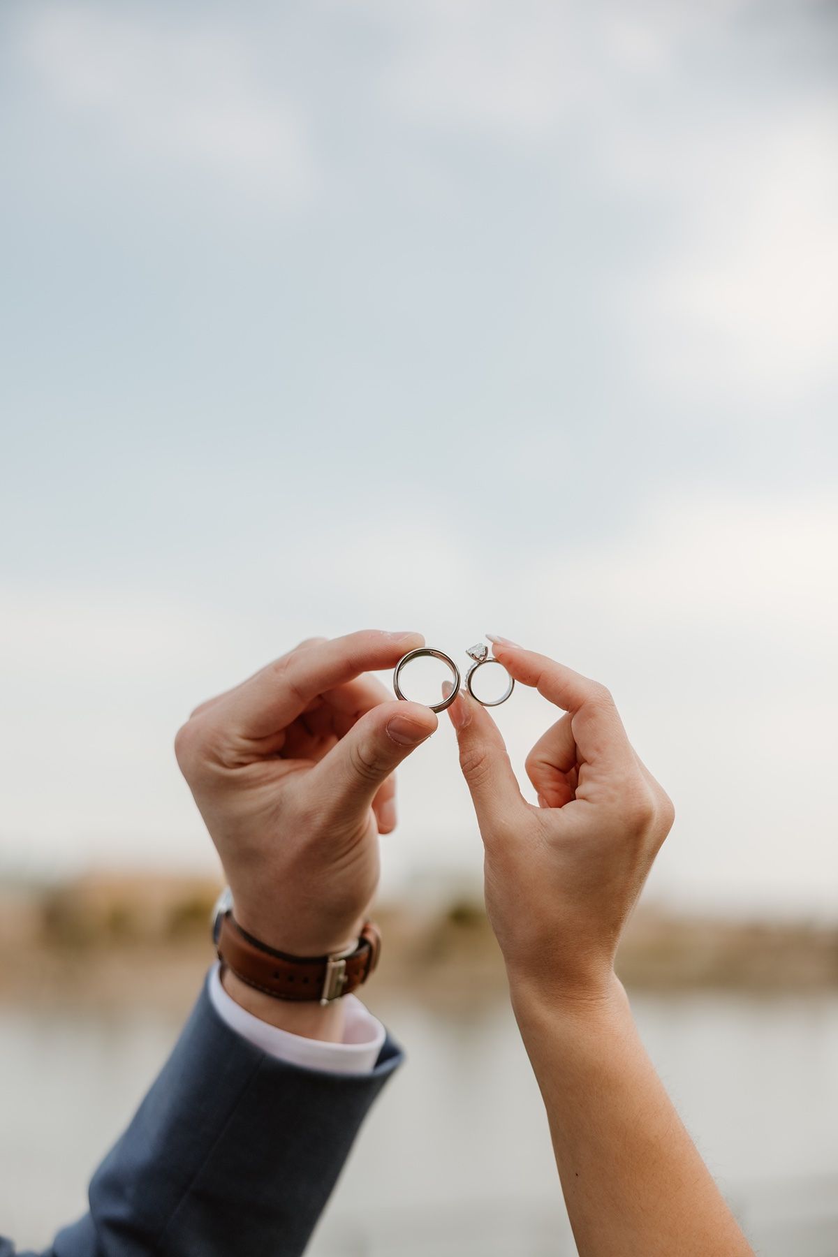 Couple holding two wedding rings together outdoors near water, groom wearing blue suit and brown leather watch.