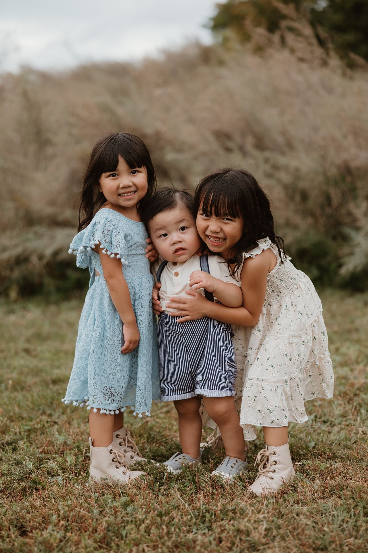 Three young children pose together in a grassy field, two girls in dresses hugging a toddler boy.