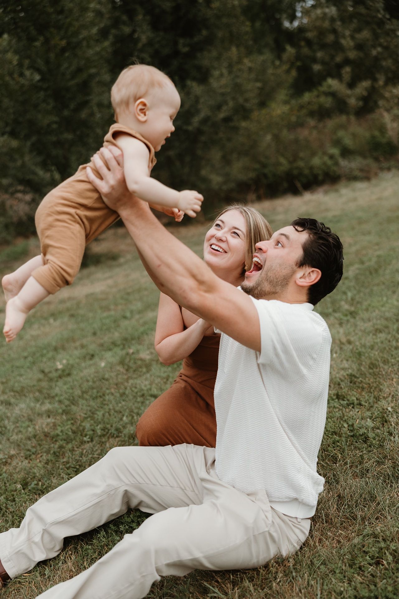 A joyful couple sits on grass, lifting their smiling baby in the air during a sunny outdoor family photo session.