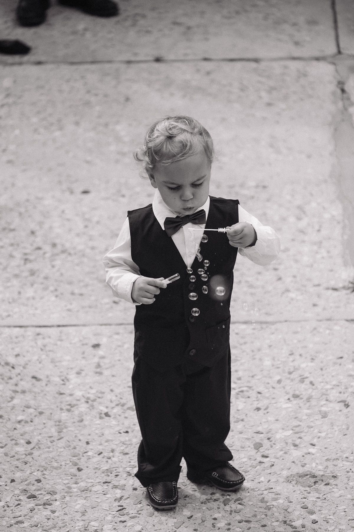 Adorable toddler boy in formal suit and bow tie blowing bubbles outdoors in black and white photo.