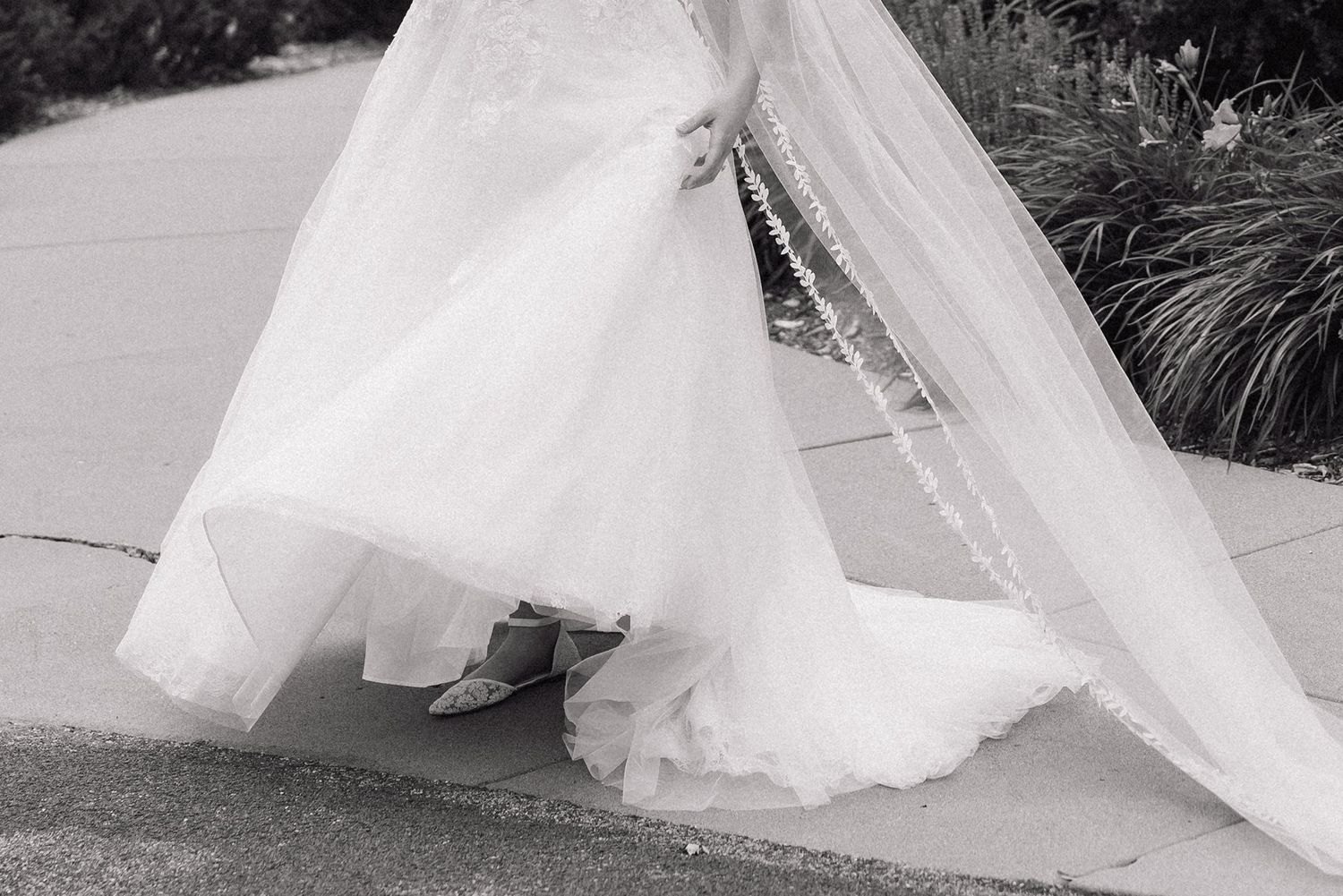 A wedding bouquet of white and blue flowers held between a black tuxedo and white wedding gown on a paved road.