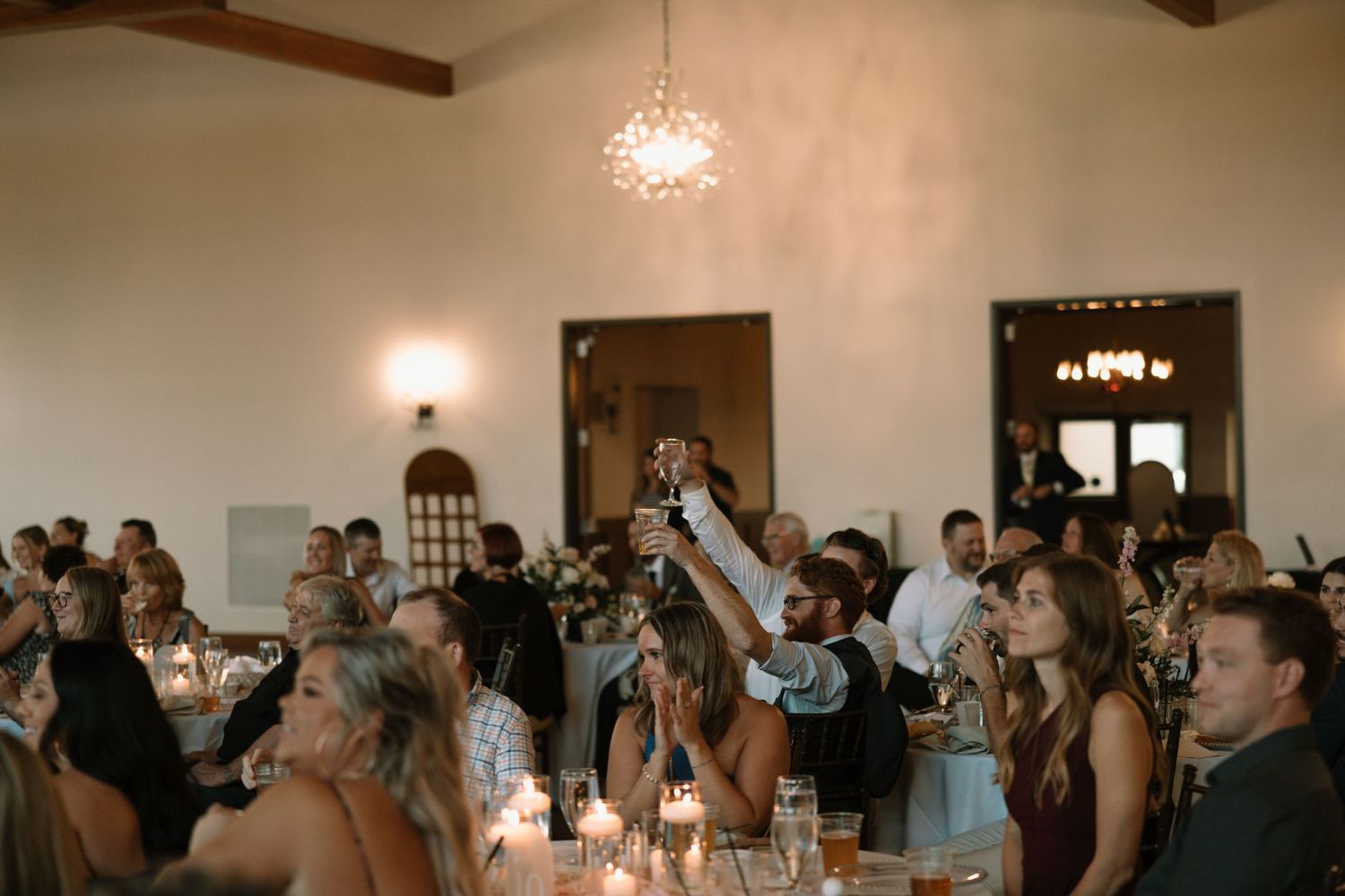 Wedding reception guests raise their glasses in celebration during a toast at an elegant venue with vaulted ceilings.