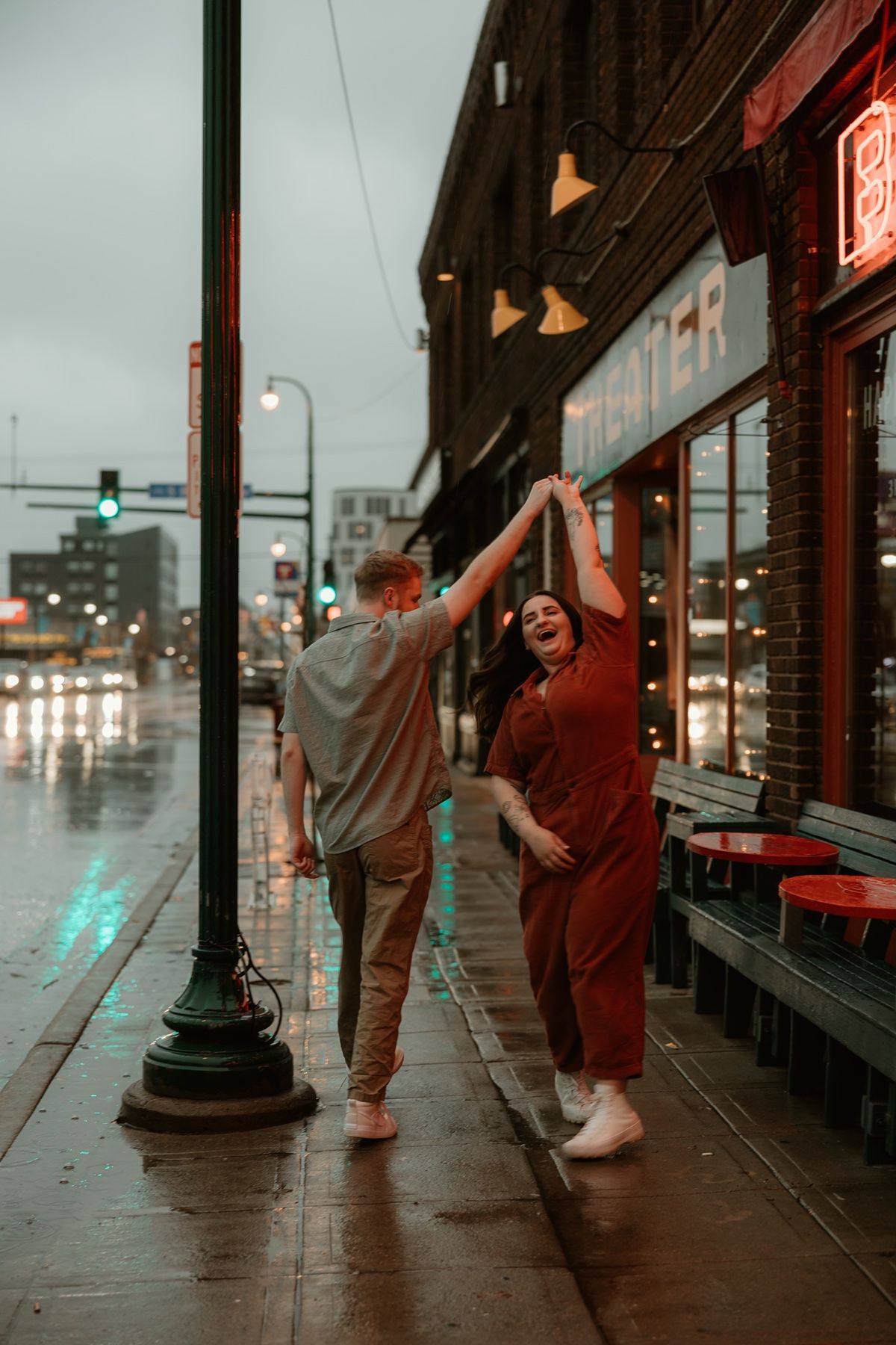 A couple dances playfully in the rain on a wet sidewalk at night under storefront lights.