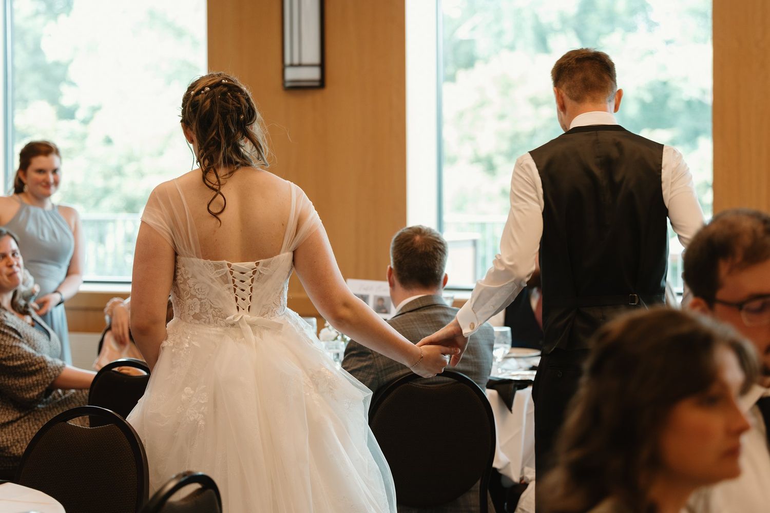 A bride and groom mingle with wedding reception guests in a banquet room with large windows at sunset.