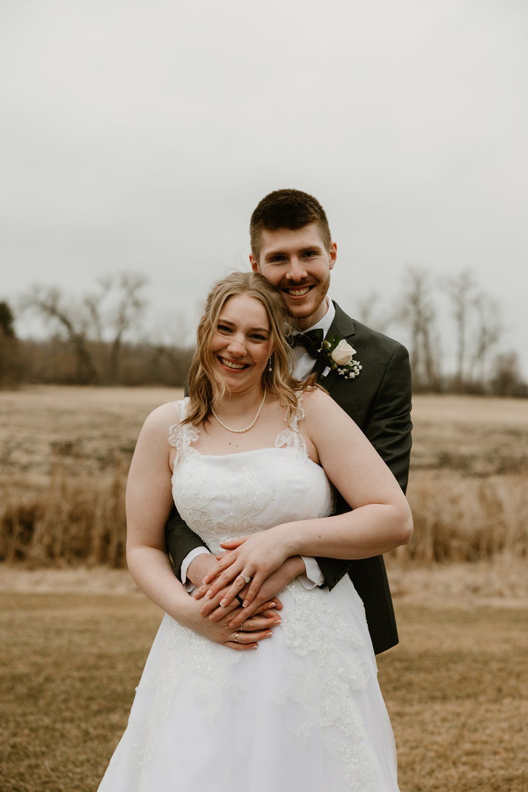 A bride and groom pose together outdoors in a rustic field setting during their wedding photoshoot.