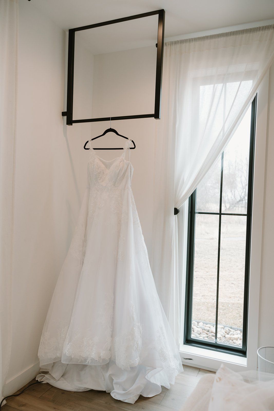 A white wedding dress hangs elegantly against a white wall next to a tall window with sheer curtains.